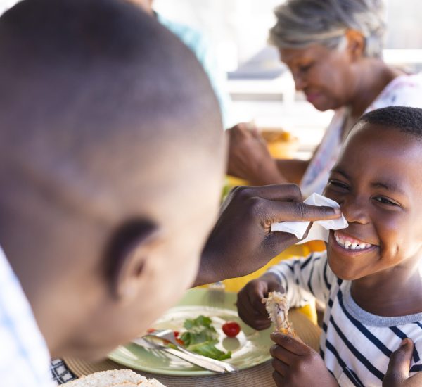 Multiracial father cleaning smiling son's nose with tissue paper while having lunch at dining table. Family, food, care, unaltered, togetherness, childhood and home concept.