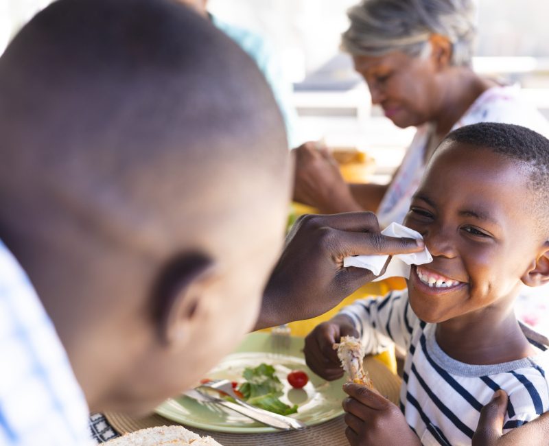 Multiracial father cleaning smiling son's nose with tissue paper while having lunch at dining table. Family, food, care, unaltered, togetherness, childhood and home concept.